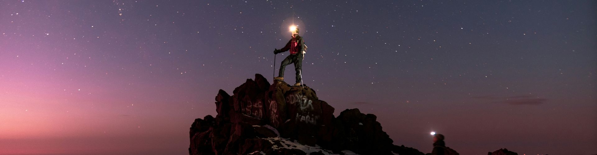 Bergsteiger mit Stirnlampe auf dem Gipfel des Jebel Toubkal bei Nacht unter klarem Sternenhimmel.