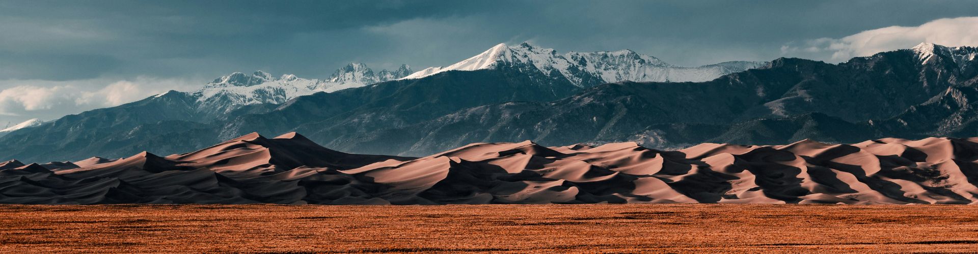 Marokko Wüstentour – Kamel in der Sahara bei Sonnenuntergang Sanddünen der Wüste Gobi vor schneebedeckten Bergen unter dramatischem Himmel