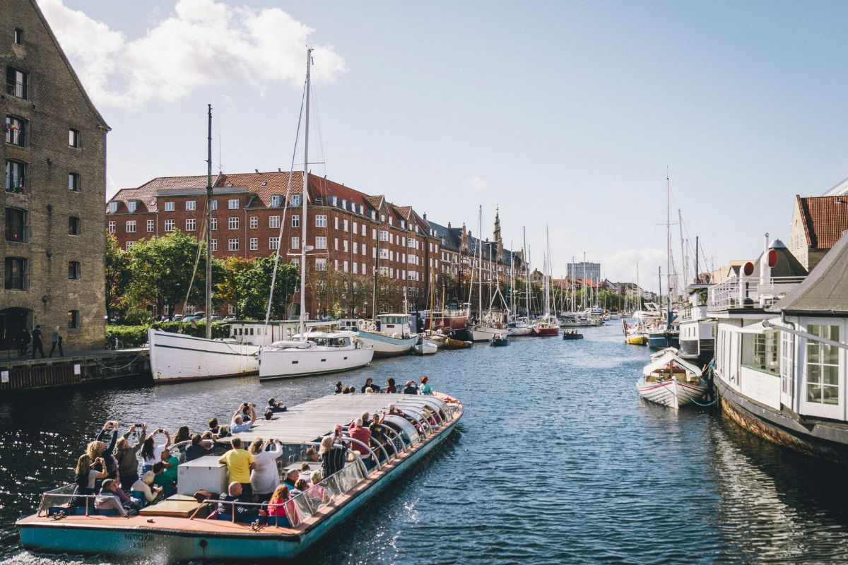 Gemütlich schippern, Kamera zücken, Sehenswürdigkeiten auf Ansage. Standardprogramm mit Anschnallgarantie. Touristen auf einer geführten Hafenrundfahrt in einer europäischen Stadt mit Ziegelhäusern und Segelbooten