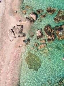 Rosa schimmernder Sandstrand von Elafonissi mit Felsen im türkisfarbenen Wasser, aufgenommen aus der Luft.