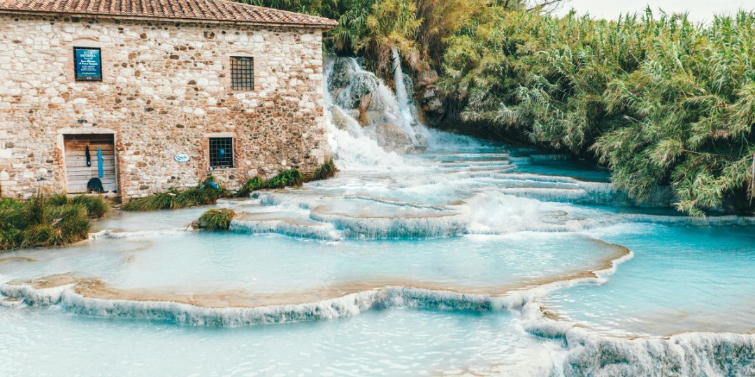 Natürliche Thermalbecken der Cascate del Mulino in Saturnia, Italien, mit kristallklarem, türkisfarbenem Wasser und einem historischen Steingebäude.