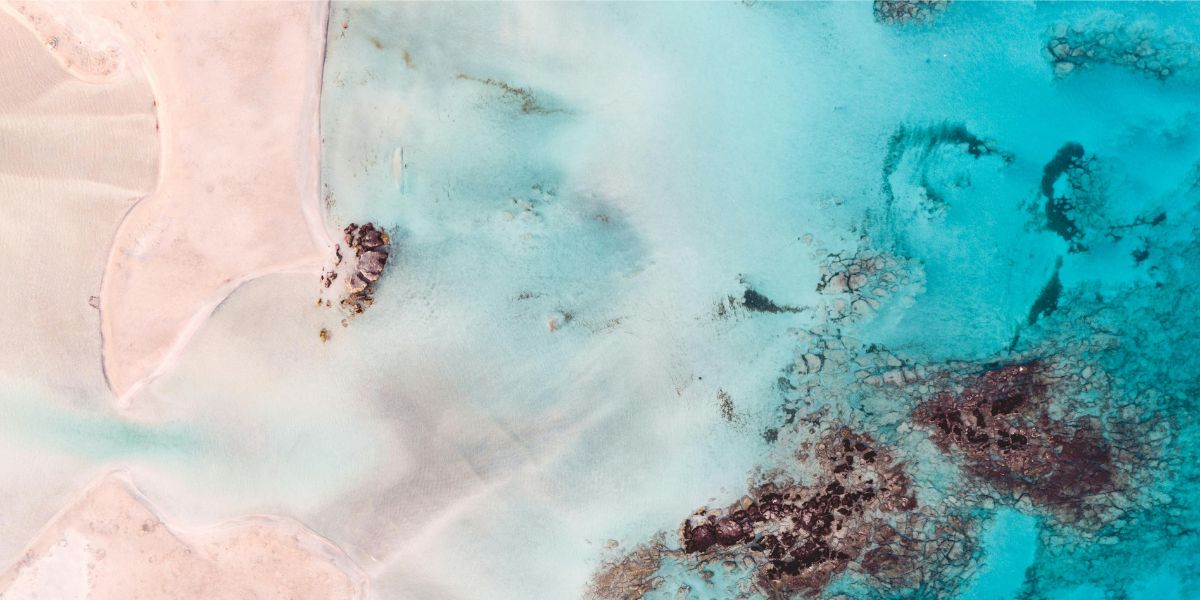 Luftaufnahme des Elafonissi Strands auf Kreta, Griechenland, mit rosa schimmerndem Sand und flach abfallendem türkisblauen Wasser.